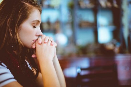A side-on photo of a girl sat at a table. She has her eyes closed, and her hands together resting against her mouth. She looks to be in deep thought.