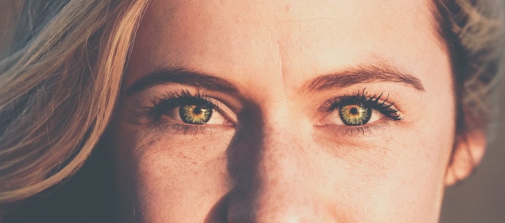 A close-up image of a woman’s face, showing her green eyes, freckled checks, and there is a suggestion of a smile.